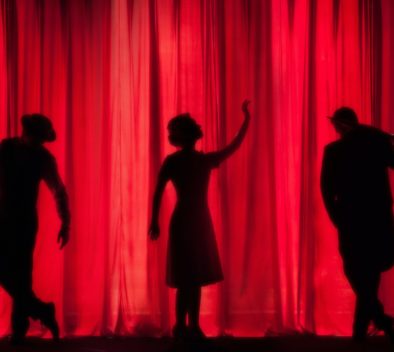 Kuvassa on punainen esirippu ja kolmen ihmisen varjokuvat, jotka esiintyvät teatterinäyttämöllä.